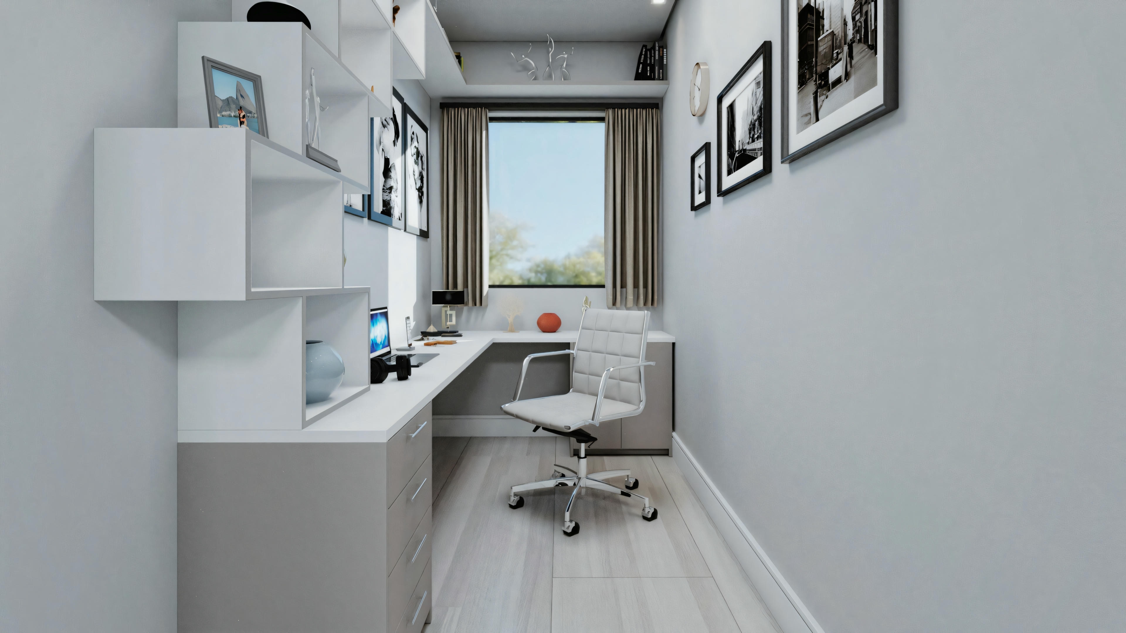 A narrow home office with white walls, a desk with a computer and chair, shelves, and pictures on the wall. There is a window in the background allowing natural light and a view of outdoor greenery.