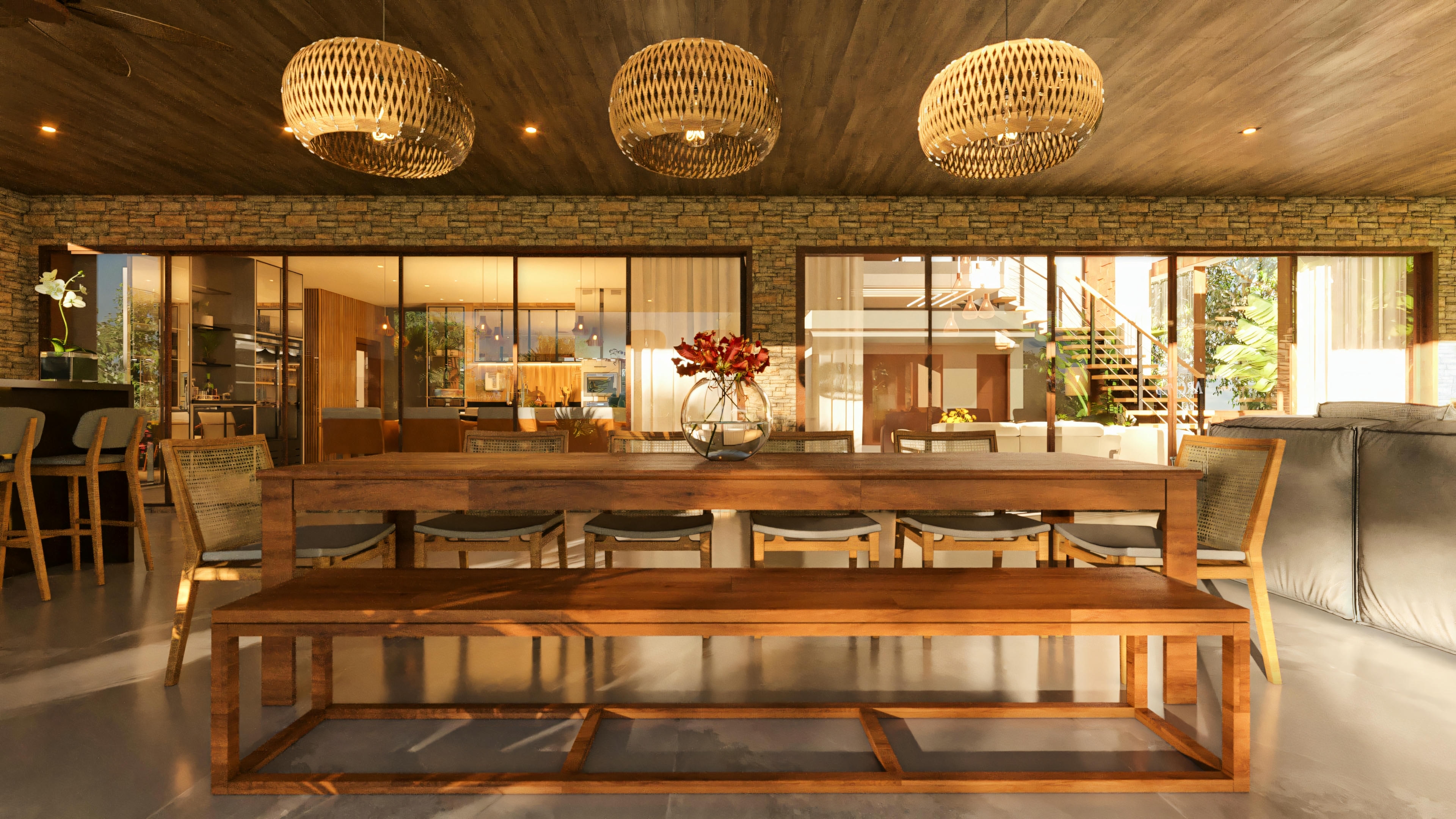 Terraza gourmet de casa de lujo con amplia mesa de comedor y banco de madera, bajo lámparas colgantes de fibra natural, con pared de piedra al fondo y grandes puertas de vidrio que integran al interior de la residencia.