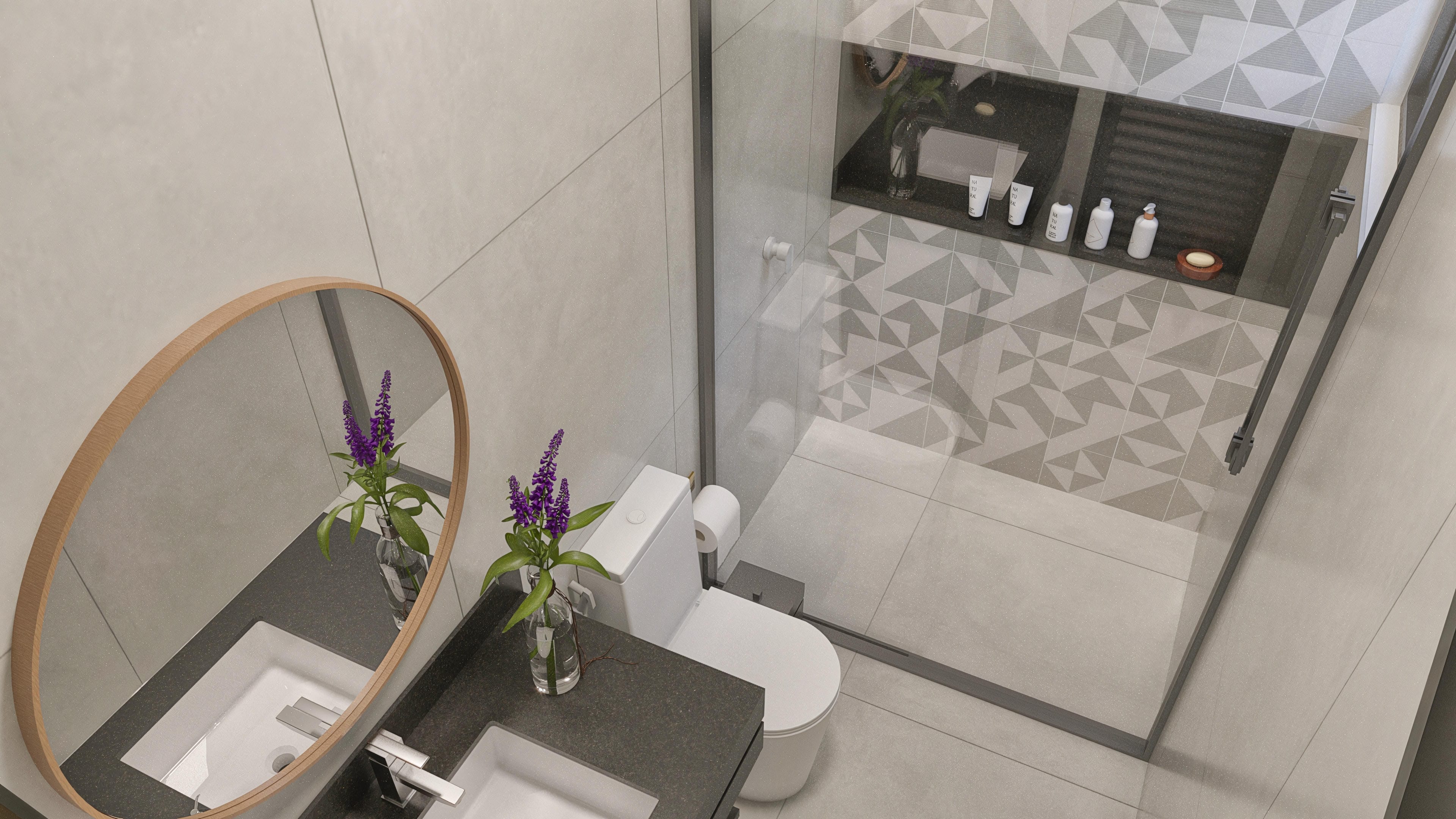 Interior de un baño moderno con encimera de granito oscuro, lavabo sobre encimera y espejo ovalado con marco de madera. La ducha de cristal templado revela azulejos con patrón geométrico en la pared.