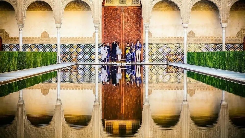 Um pátio da Alhambra, na Espanha, com um espelho d'água central e arquitetura islâmica detalhada ao fundo.