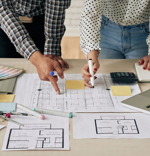 Two people, one in a plaid shirt and the other in a polka dot blouse, leaning over a floor plan with various office supplies, pointing to different parts of the project. The laptop on the left is open and there is a calculator on the table.