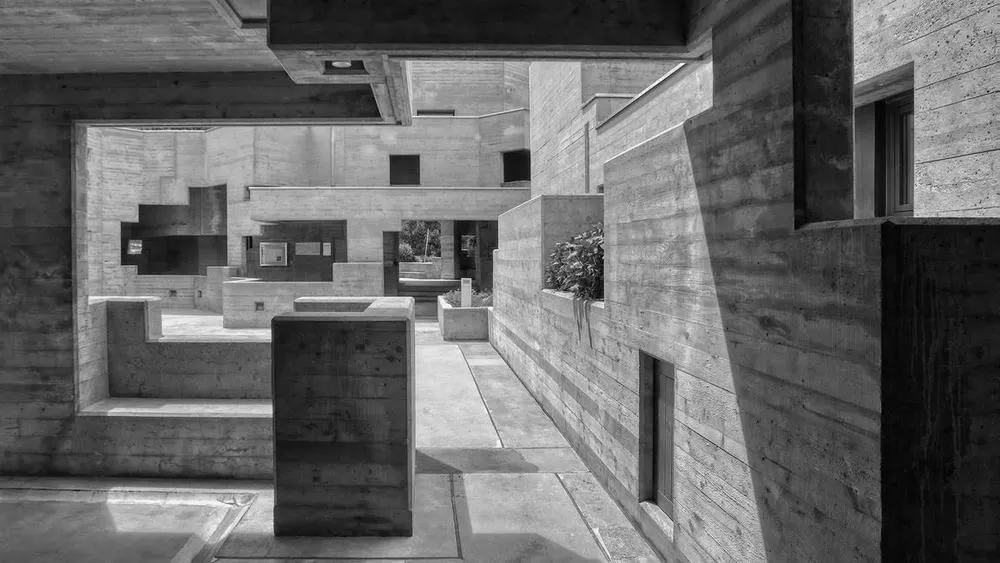 Path between concrete structures in a brutalist building, with striking shadows and natural lighting highlighting textures. Black and white image.
