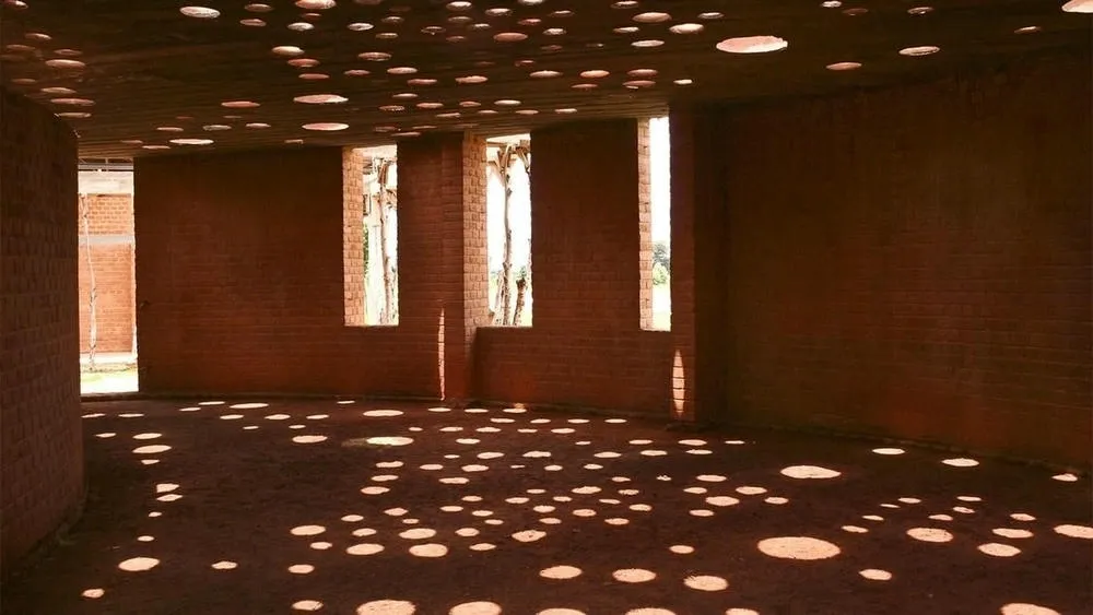 Interior de uma construção com paredes de tijolos e buracos redondos no teto e nas paredes, criando padrões de luz e sombra no chão.