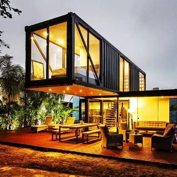 Casa hecha de contenedores negros y grandes ventanas, con un área exterior iluminada y amueblada para ocio, por la noche.