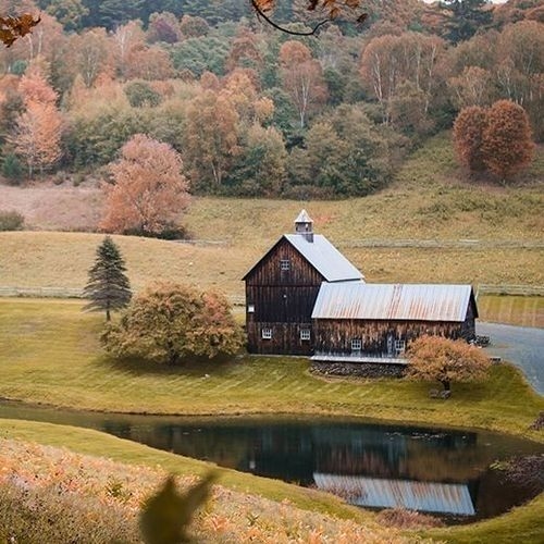 Una encantadora casa de campo de madera oscura con techo gris, rodeada de árboles con follaje de otoño y un pequeño lago que refleja la estructura.