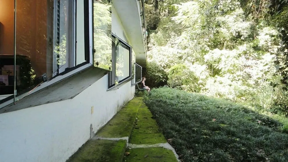 Lateral de uma casa com grandes janelas e vegetação rasteira no telhado e na lateral. Uma pessoa está sentada em uma cadeira na varanda, observando a paisagem natural. O projeto busca integrar o ambiente interno com o exterior.