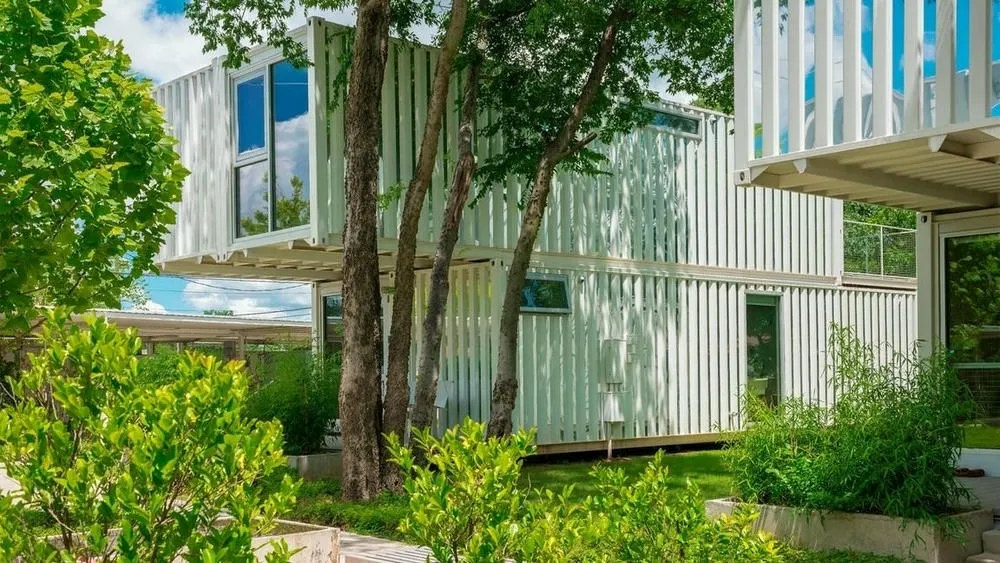 Dos casas hechas de contenedores blancos, con grandes ventanas y balcones, rodeadas de vegetación y árboles en un césped.