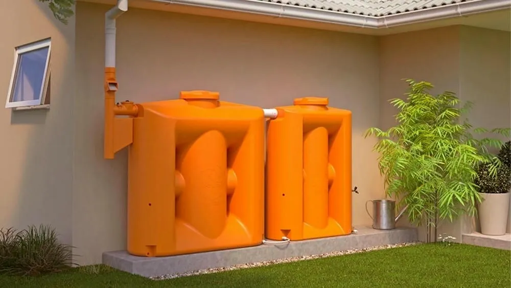 Two large orange cisterns installed next to a house with a roof, collecting water from the gutter. Around, lawn and plants.