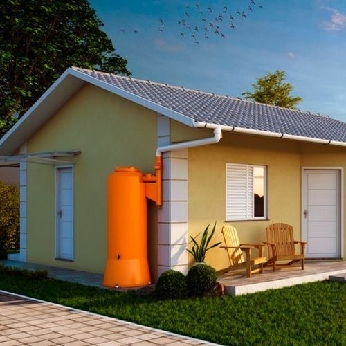 Casa amarela com cisterna laranja para captação de água da chuva. Há um jardim ao redor com cadeiras de madeira e vegetação.