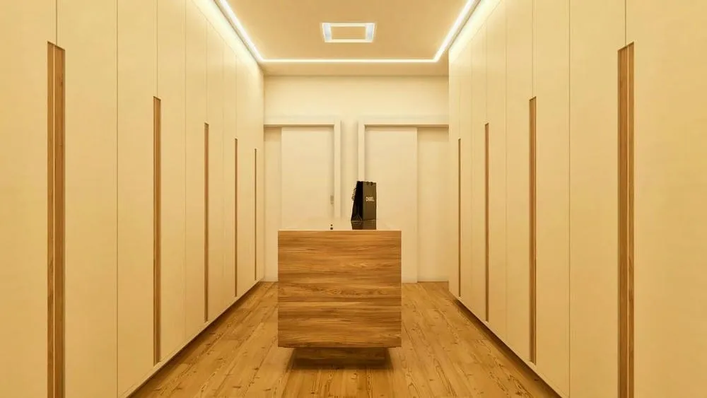 Corredor de closet com armários fechados em tons claros e iluminação embutida, com uma ilha central de madeira.