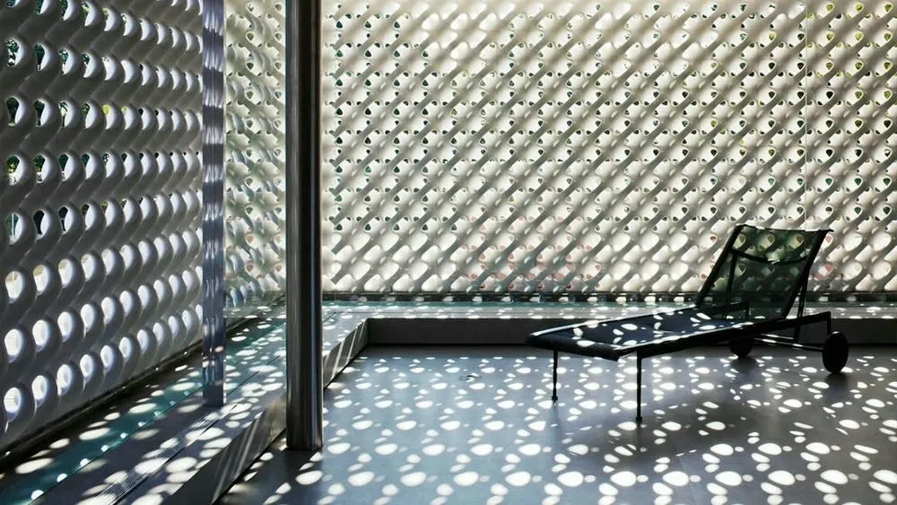Interior environment with a white cobogó wall creating a play of light and shadow on the floor, with a lounge chair next to it. The light filtered by the cobogós creates an artistic effect in the space.