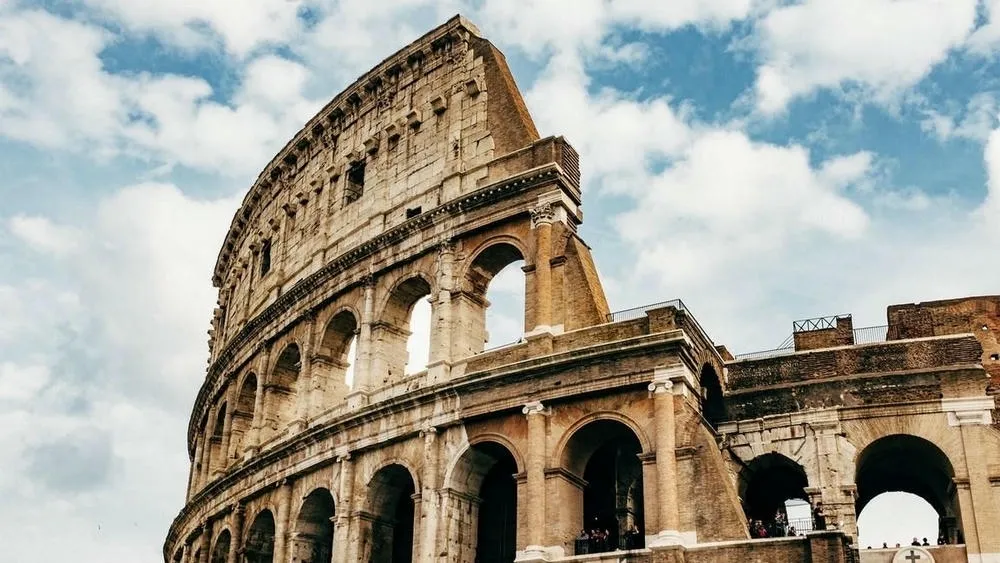 Parte da fachada do Coliseu em Roma, Itália, vista de baixo para cima em um dia ensolarado com nuvens brancas. A estrutura antiga de pedra e arcos é proeminente, mostrando a imponência da arquitetura clássica romana.