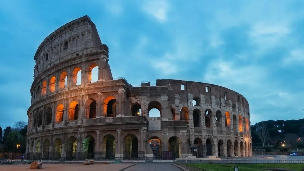 Vista noturna do Coliseu em Roma, Itália, com suas arcadas iluminadas em tons quentes sob um céu azul-escuro com nuvens. A imponente estrutura de pedra mostra a escala e a beleza da arquitetura clássica romana.
