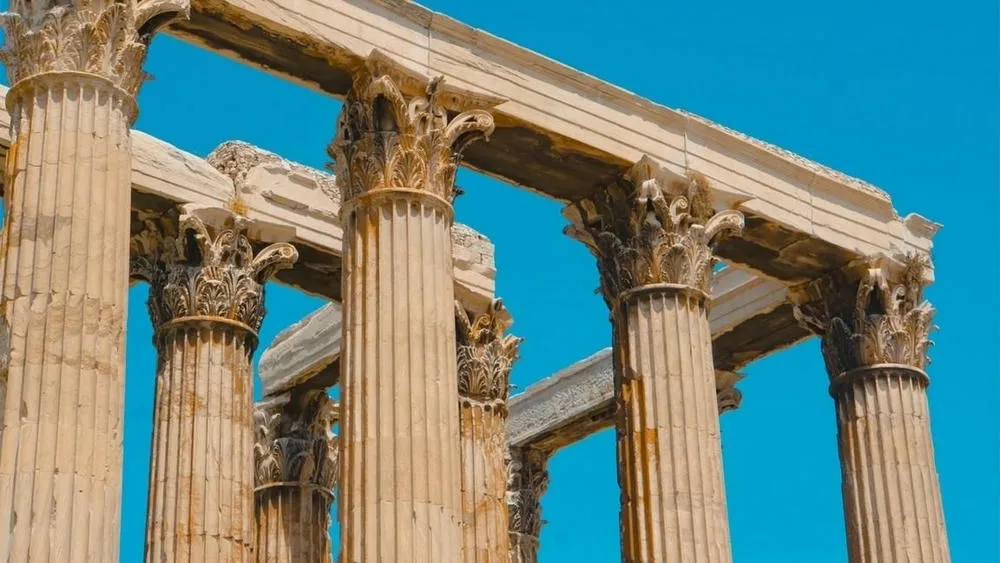 Detalhe de colunas coríntias em um templo antigo, com o céu azul vibrante ao fundo. Os capitéis são ricamente decorados com folhas de acanto, exemplificando a ornamentação característica da arquitetura clássica.