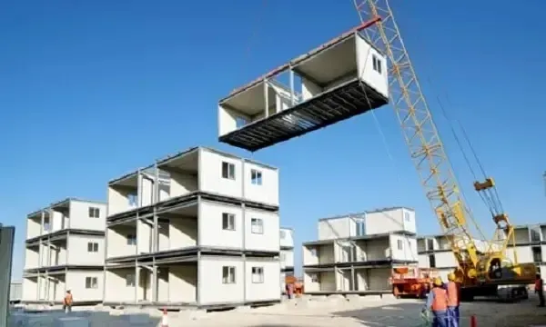 Módulos de casas siendo izados por una grúa en un sitio de construcción, con otros módulos ya montados al fondo, bajo un cielo azul.