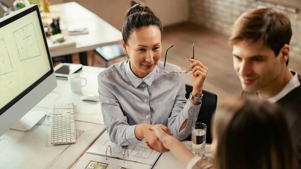 Una arquitecta sonriente, sosteniendo gafas, saluda a una cliente sobre una mesa con planos de proyectos.