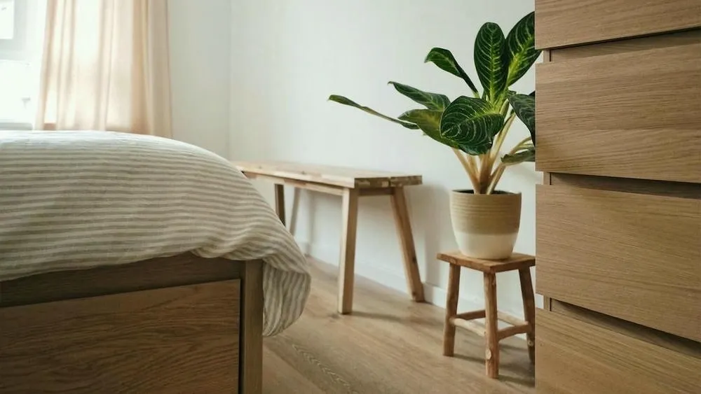 Um canto de quarto minimalista com uma planta grande em um vaso de dois tons sobre um pequeno banco de madeira, ao lado de uma cama com lençóis listrados e uma bancada de madeira, ressaltando a conexão do estilo escandinavo com a natureza.