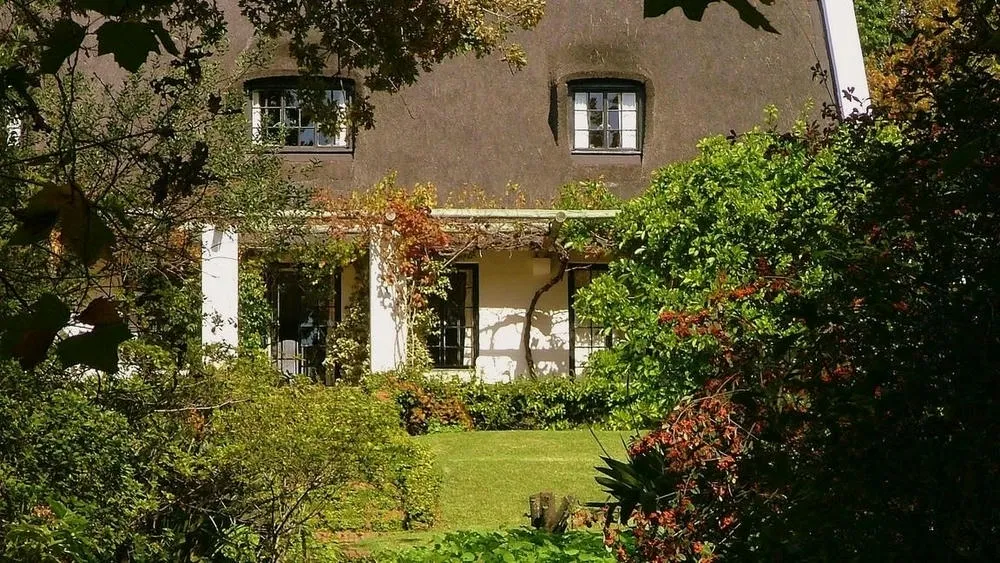 Part of a country house with a neutral-colored facade and dark roof, partially hidden by foliage and trees with autumn tones, with a porch and windows.