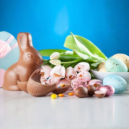 Conejo de chocolate junto a huevos de Pascua coloridos y flores en tonos pastel, con una caja de regalo azul al fondo, sobre una superficie clara y fondo azul.