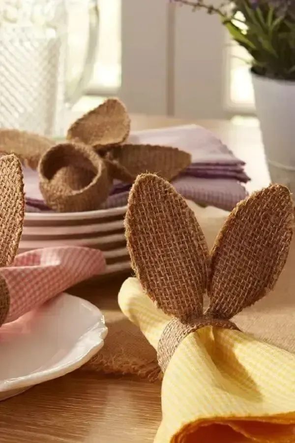 Detalhe de guardanapos de tecido com anéis em formato de orelhas de coelho de juta, ao lado de pratos brancos.