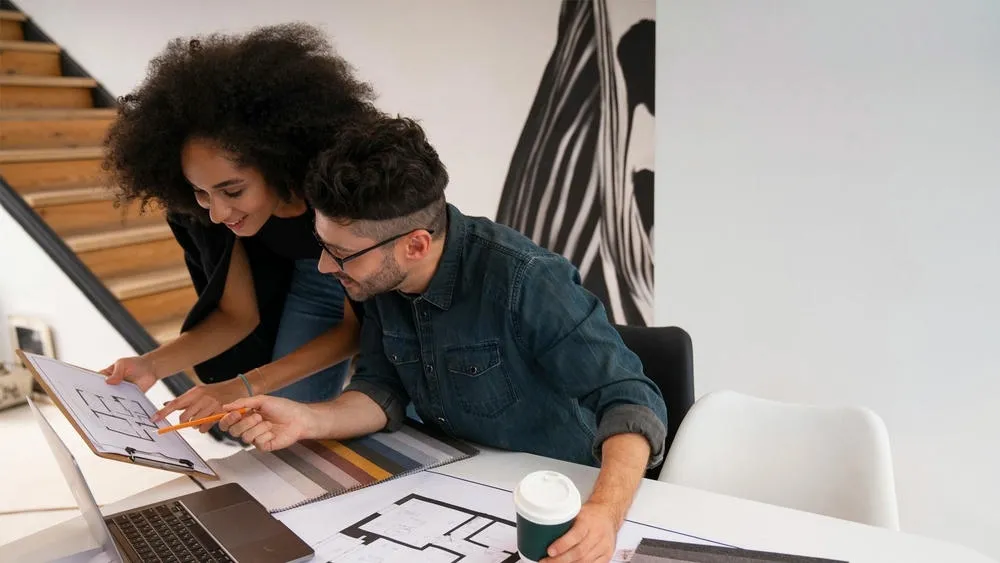 Dos personas, un hombre y una mujer, en una oficina, inclinadas sobre un plano, discutiendo y señalando el dibujo.