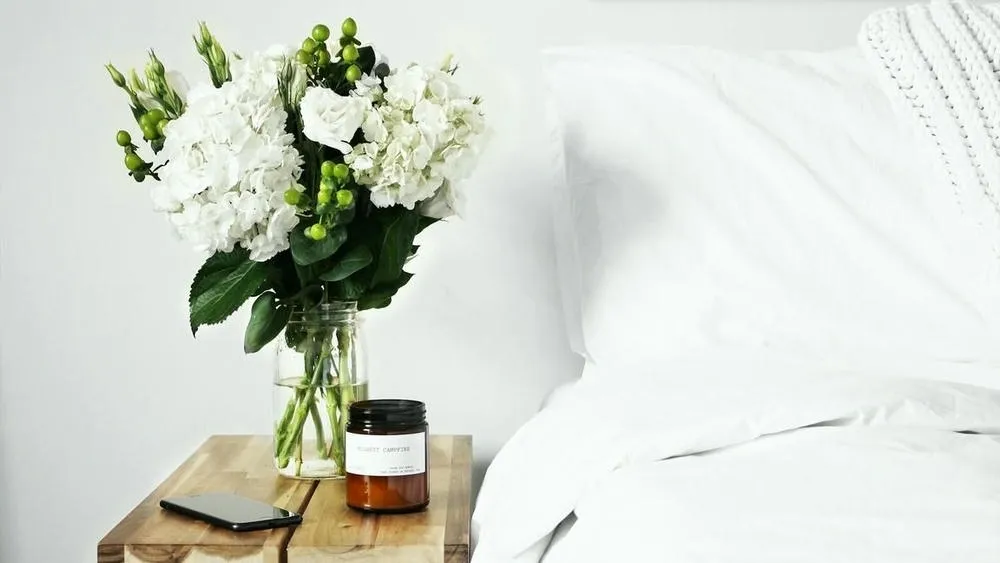 Um vaso com flores brancas e folhagens verdes em uma mesa de madeira ao lado de uma cama com lençóis brancos, um telefone celular e uma vela aromática, destacando a paleta de cores claras e os materiais naturais do estilo escandinavo.