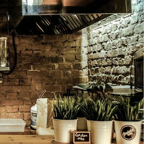Cozinha com estética industrial apresentando parede de tijolos aparentes, coifa de aço inoxidável e plantas em vasos brancos, remetendo a um ambiente rústico e moderno.