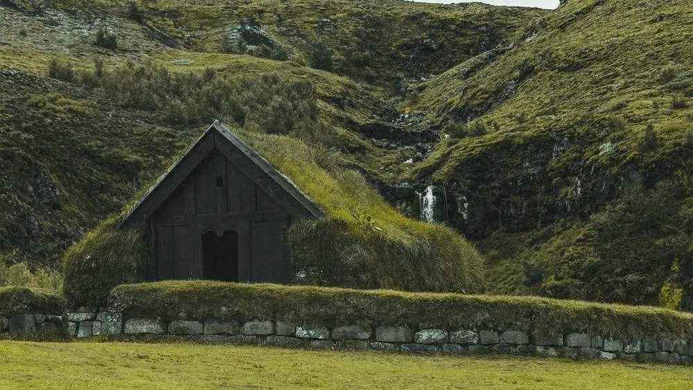 A traditional wooden house with a grass-covered roof, on a green hillside in Iceland, with a small waterfall in the background.