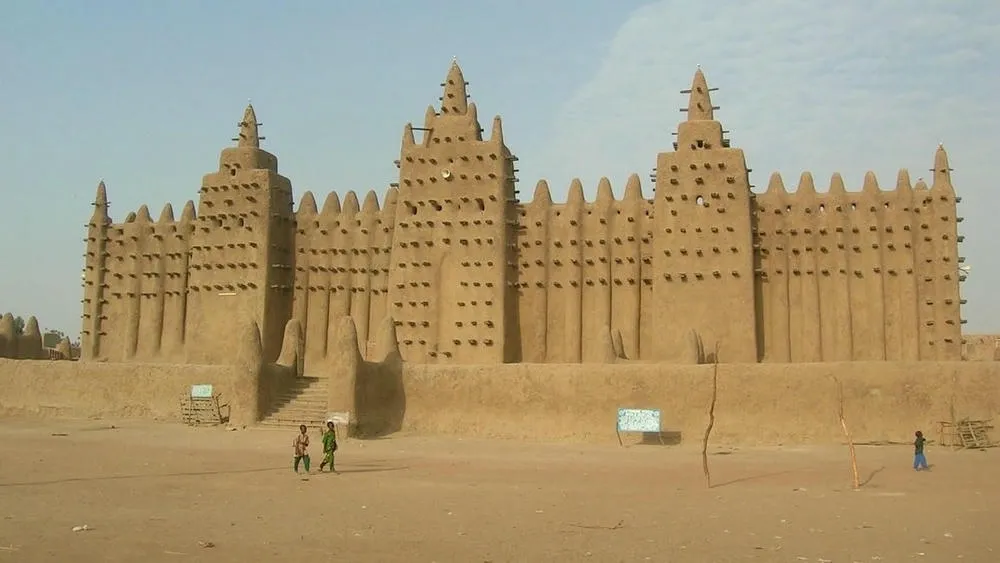 A Grande Mesquita de Djenné, uma construção de adobe com torres e detalhes salientes, no Mali, com duas pessoas caminhando em frente.
