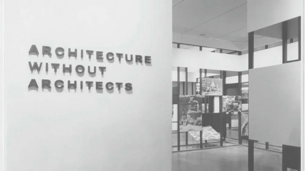 Una exposición de arte con el título 'Architecture Without Architects' en una pared, y paneles con fotos de construcciones vernáculas al lado.