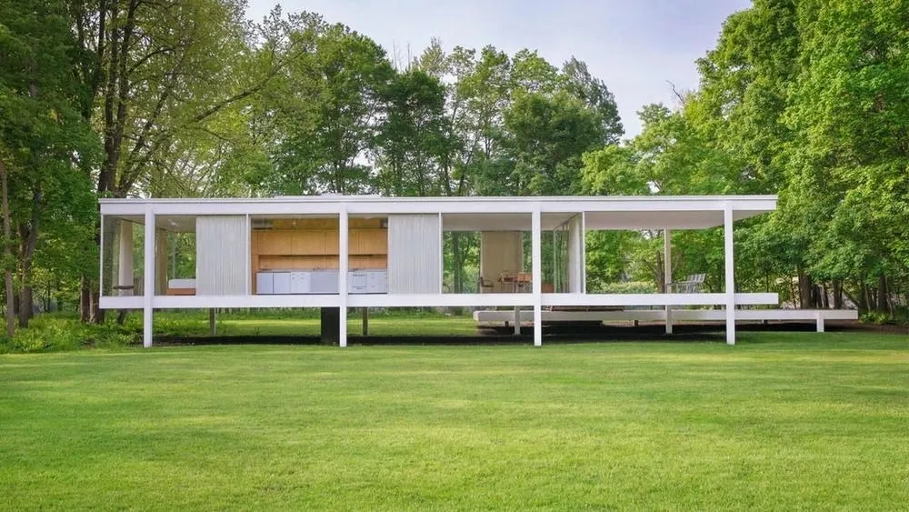 Side view of the Farnsworth House, a rectangular glass and steel structure elevated from the ground, surrounded by trees, illustrating the simplicity and connection with nature of minimalism.
