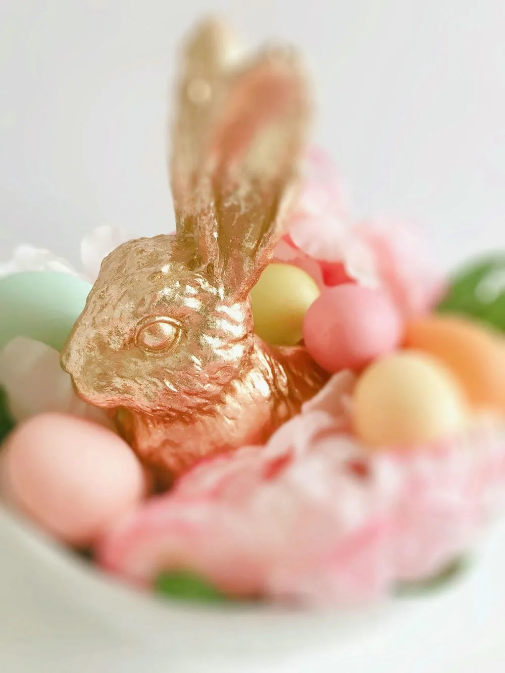 Golden chocolate bunny and colorful Easter eggs in pastel tones, surrounded by light pink fabric and blurred flowers.