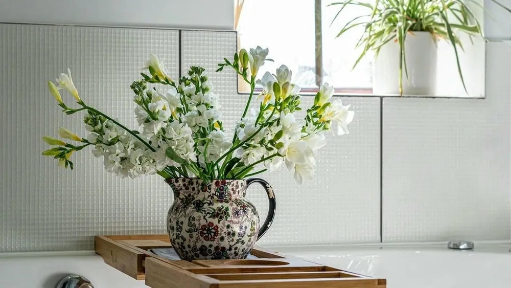 Um vaso florido com flores brancas e algumas hastes verdes, posicionado sobre uma bandeja de madeira em uma banheira. O fundo é composto por uma parede de azulejos brancos texturizados e uma janela, de onde entra luz natural. Um vaso menor com uma planta verde pode ser visto no canto superior direito.