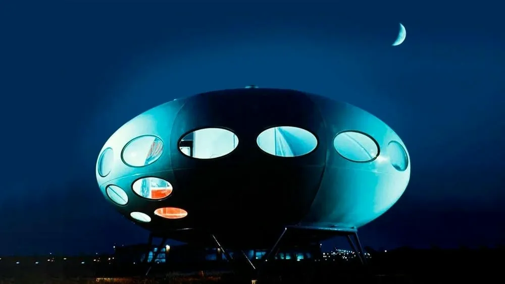 A Futuro House, uma casa com formato de disco voador, iluminada em tons de azul à noite, sob um céu escuro com uma meia-lua. A entrada está iluminada em vermelho e a grama ao redor é escura.