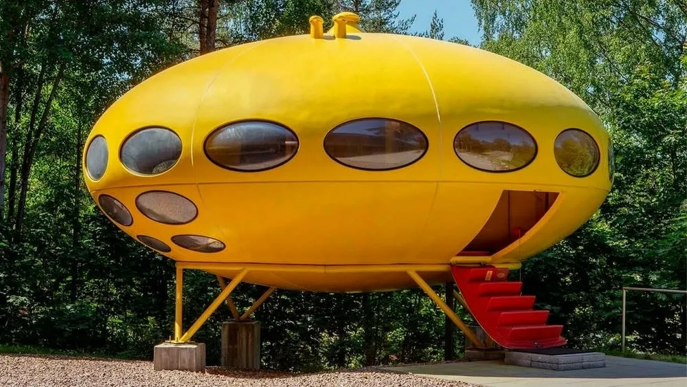 Uma Futuro House amarela, com formato de disco voador e janelas ovais, em um ambiente externo cercado por árvores. Uma escada vermelha retrátil leva à entrada principal. A casa é elevada do chão por pernas de metal.