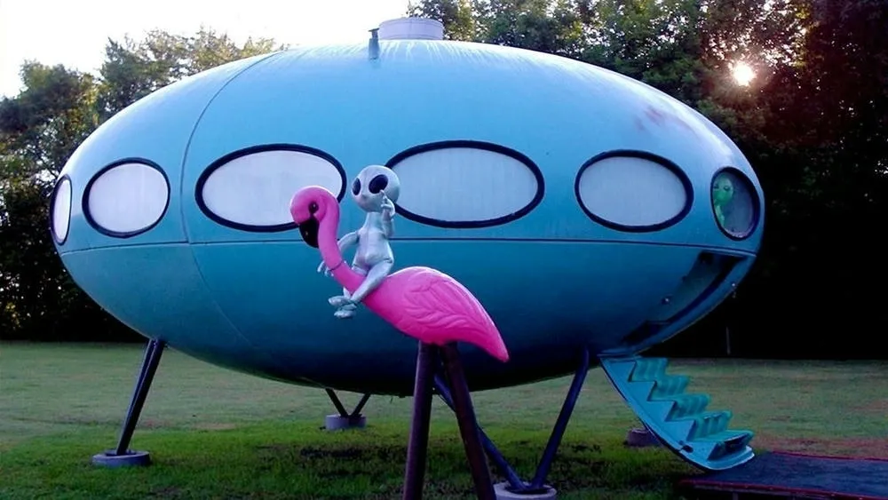 Uma Futuro House azul em um gramado, com um alienígena cinza sentado em um flamingo rosa em primeiro plano. A casa possui janelas ovais e uma escada retrátil verde.