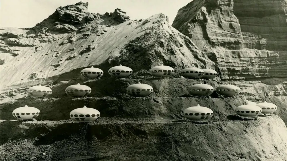 Múltiplas unidades da Futuro House, casas em formato de disco voador, dispostas em diferentes níveis de uma paisagem montanhosa e árida. O ambiente remete a um cenário desértico ou lunar, com as casas futuristas se destacando contra as rochas.