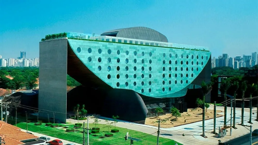 Vista externa angular do Hotel Unique em São Paulo, um edifício de arquitetura contemporânea com fachada curva em tom de azul esverdeado e várias janelas circulares. A estrutura se eleva sobre pilares, com áreas verdes ao redor e edifícios ao fundo.