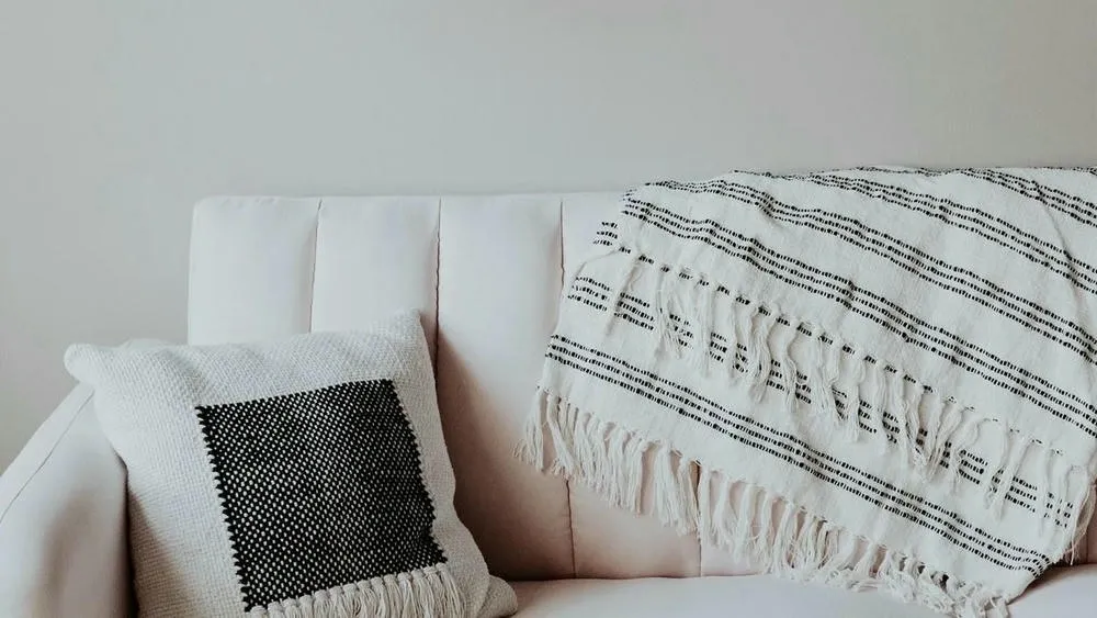 Um detalhe de um sofá claro com uma almofada branca e preta texturizada e uma manta listrada, transmitindo a sensação de aconchego e conforto do conceito Hygge no design escandinavo.