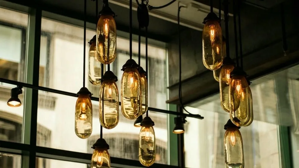 Luminárias pendentes de metal com lâmpadas expostas em um ambiente com teto alto e parede de tijolos, representando a iluminação no estilo industrial.