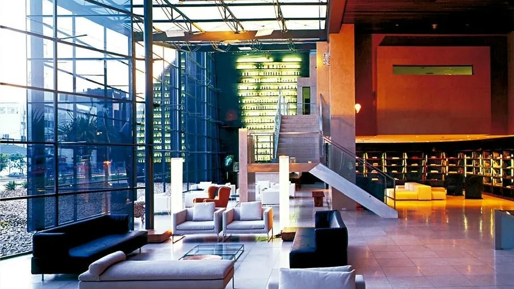 Interior de um grande lobby com pé direito alto, grandes janelas de vidro e muita luz natural. Há sofás e poltronas em tons claros, e prateleiras iluminadas ao fundo, criando um ambiente amplo e sofisticado.