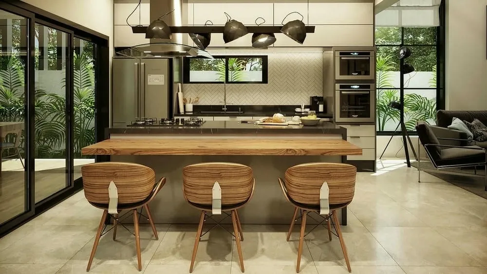 Cocina con encimera negra y sillas de madera, destacando lámparas colgantes de metal en estilo industrial. Al fondo, ventana con vista al verde.