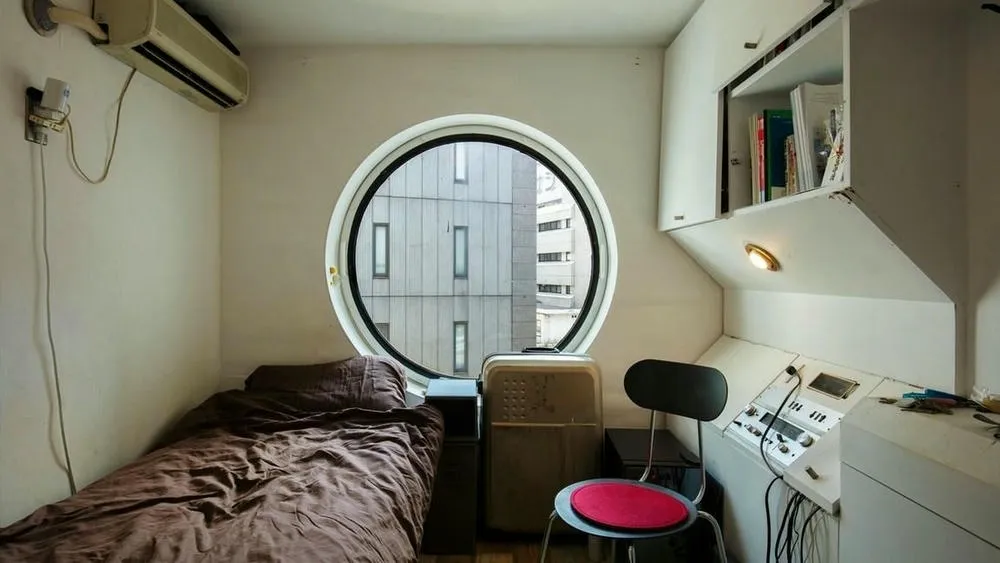 Interior de una pequeña cápsula modular con una cama, una ventana redonda y una pequeña estación de trabajo con equipos electrónicos.