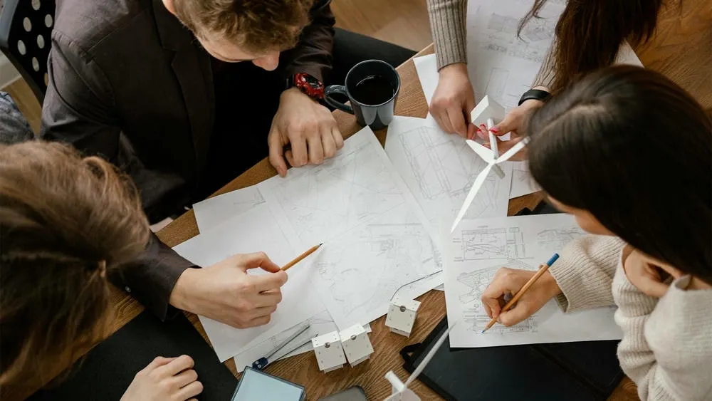 People at a table reviewing plans and models, with pencils and other design materials.