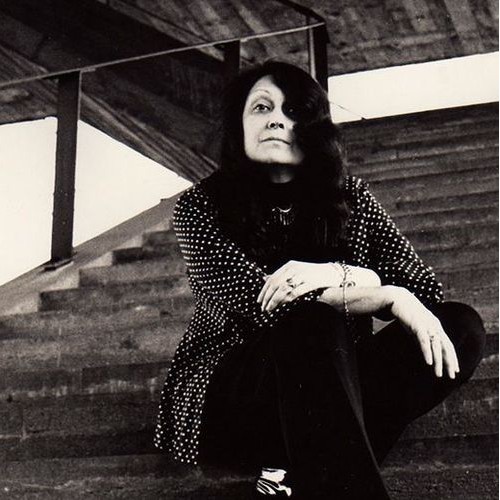 Retrato em preto e branco de Lina Bo Bardi sentada em degraus de concreto com estrutura aparente, destacando sua presença icônica na arquitetura brasileira.