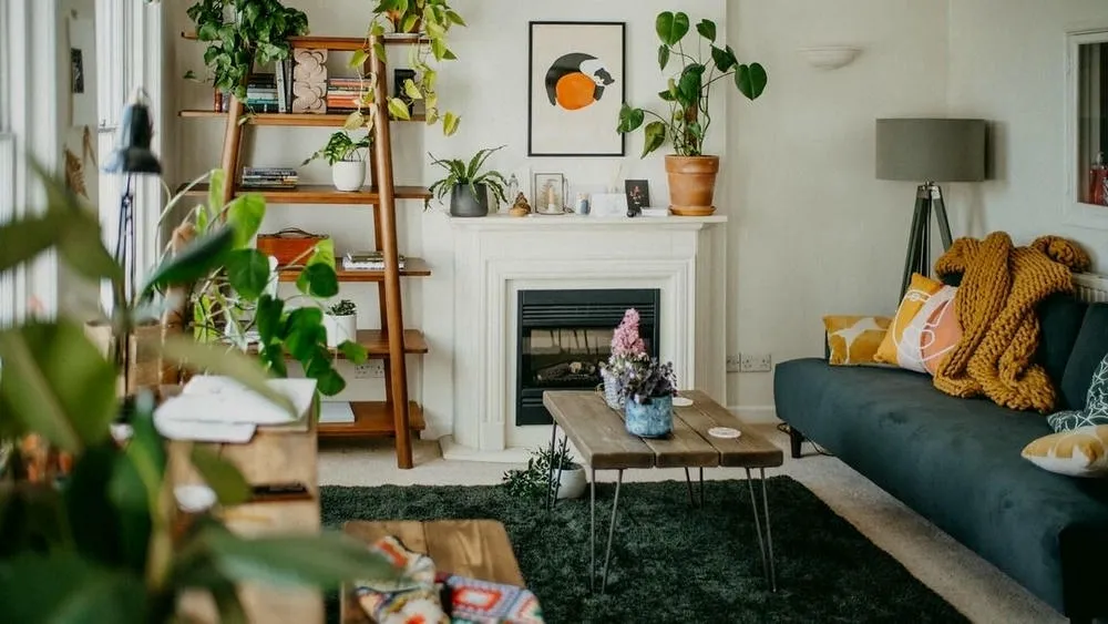 A living room decorated with various plants, including a large plant in a floor pot, hanging plants on a wooden shelf, and smaller plants on the fireplace. A gray sofa with colorful cushions, a wooden coffee table, and a white fireplace complete the setting. Natural light enters through the side window, creating a cozy and lively space.