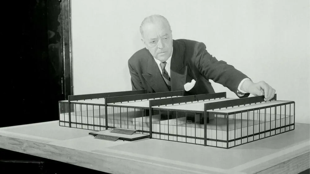 Ludwig Mies van der Rohe analyzing an architectural model of a project with straight lines and large glass panels, characteristic of the minimalist style.