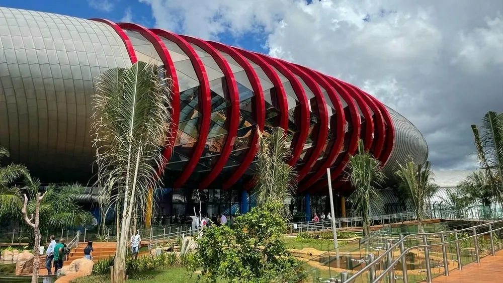 Um edifício com fachada curva, predominante em tons de cinza e com grandes anéis vermelhos e vidros que se projetam para fora. Palmeiras e vegetação complementam a paisagem em frente ao edifício. O edifício possui um design moderno e inovador, com materiais tecnológicos.