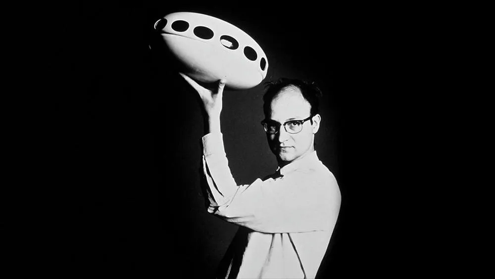 Retrato em preto e branco do arquiteto Matti Suuronen, que segura uma miniatura da Futuro House acima de sua cabeça. Ele usa óculos e olha diretamente para a câmera.