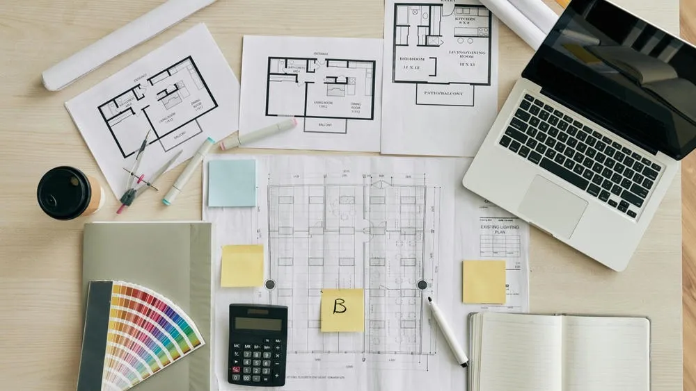 Vista superior de una mesa de oficina con varios planos bajos de arquitectura, un portátil abierto, calculadora, bolígrafos de colores, notas adhesivas y un libro de muestras de colores. Una mano está señalando uno de los planos.